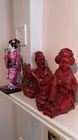 Photo of two red resin Asian figurines and a pink kimono doll placed on a mantelpiece, showing detailed features and traditional clothing.