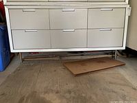 Front view of white six-drawer metal cabinet on metal legs showing drawer arrangement and handle design.