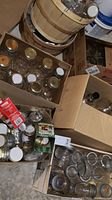 Multiple boxes and a wooden bushel basket filled with a variety of glass canning jars, lids, and recycled food jars.