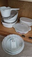 Shelf display of white glass bakeware and cookware including pans with lids and small bowls.