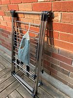Folding chrome drying rack leaning against brick wall with cloth bag attached.