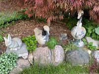 Five concrete garden animal statues in a garden setting under a tree. Visible animals include two geese, a rabbit, and two frogs among rocks and plants.