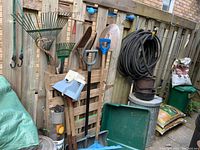 View of garden rakes, shovels, soaker hose on reel, planting tray, bags of mulch and manure, green plastic bin, plastic and galvanized cans, and bird feeder hung on wood fence.