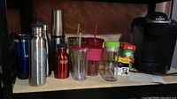 Photo of Keurig coffee machine, various coffee pods boxes, Melitta coffee filters box, and multiple stainless steel and plastic beverage bottles and tumblers in assorted colors arranged on a shelf.