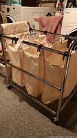 Photo showing front and side view of laundry sorter with three cream-colored polycotton bags and chrome frame on wheels in laundry room.