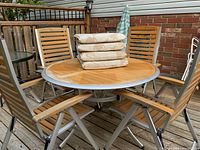 Full view of table with four wood slat reclining chairs with cushions on a wooden deck, stacked cushions on table, umbrella visible to side