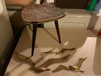 Photo showing the mid century triangular stool with mosaic tiled top and three wooden legs next to three brass birds of varying sizes.