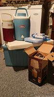 Overview photo of lot showing two Coleman coolers, one red and white Coleman beverage jug with metal closer, five blue and white ice packs, and box for Ray-O-Vac portable table lamp.