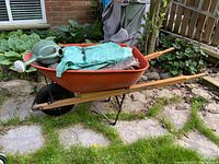 The entire lot arranged in a garden setting showing the red metal wheelbarrow with wooden handles loaded with tarps and the green watering jug on top.