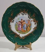 Full view of the 7-inch Limoges hand painted plate standing on wooden display. Shows the green and gold border and central classical scene.