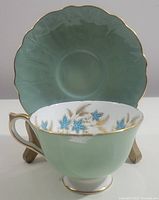 Front left side view of wide mouth tea cup with pale green and gold leaf design, showing floral interior and matching saucer behind it.