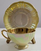 Tea cup with gold interior and iridescent pale green exterior placed in front of matching saucer with gold and iridescent finish.