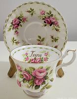 Tea cup front view showing pink rose design, gold handle, 'June' inscription inside, set against matching saucer with floral pattern and scalloped edge.