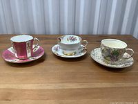 Three gold-accented fine china tea cups and matching saucers arranged side by side, clearly showing their design details and colors.