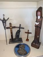 Wide view showing all 8 devotional religious items arranged spaced on shelf: 5 crucifixes, 1 statue, 1 bas-relief medallion, and small figurines.