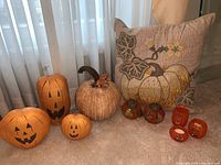 Photo showing full lot arranged on floor: Pottery Barn pillow, three stuffed orange pumpkins, resin pumpkin with metal leaves, two glass pumpkins with metal tops, and three small glass votive jack-o-lanterns.
