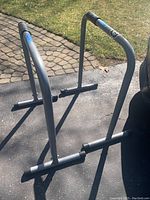 Pair of gray metal dip bars positioned outdoors on pavement with grass background, showing front and side angles with foam padded hand grips visible.