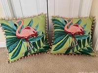 Two square outdoor pillows with flamingo and tropical leaf print, pom-pom ball fringe on edges, posed on beige carpet.