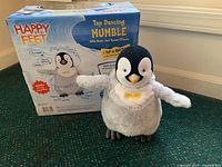Front view of the Mumble plush toy positioned in front of its original Happy Feet branded box. Shows the plush penguin's body and details clearly.