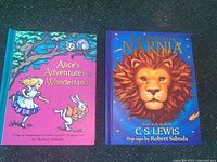 Two pop-up books by Robert Sabuda side by side on a dark surface: Alice's Adventures in Wonderland and The Chronicles of Narnia.