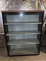 Front view of black steel bookcase cabinet with wood grain laminated top and sliding glass doors revealing four shelves with contact paper lining.