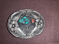 Front view of the oval silver belt buckle showing carved floral and leaf details with turquoise and coral stones.