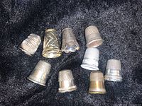 Top-down image showing all nine vintage thimbles arranged on a dark surface showing different materials and decorative designs.