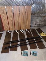 Photo shows a set of dark and light wood laminate shelves, metal brackets in white and brown, black metal racks, and two finials arranged on the floor against a stone wall.