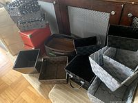 Photo showing multiple woven baskets in black, gray, and brown, red metal storage boxes, and hanging gray metal baskets arranged on and beside chairs.