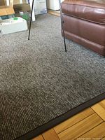 Close-up photo showing the textured charcoal grey pattern of the polypropylene area rug, including edge binding and part of surrounding floor and furniture.