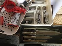 Photo showing knife set in case, stainless steel cutlery in white tray, red measuring cups, metal grater and other utensils.