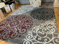 Photo of grey area rug with four large circular floral motifs in red, black, and white on a grey background, laid on a hardwood floor under diffused light.