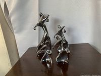 Two taller and two smaller shiny metallic abstract sculptures shown from front, placed on dark wood surface against light textured background.