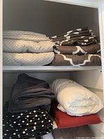 Two shelves with neatly folded bedding including patterned sheets in gray with white polka dots, dark brown with white geometric shapes, and solid dark gray, white, red, and black with white polka dots sheets or pillowcases.