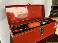 Open red metal tool box showing various hand tools inside like screwdrivers with orange, black and red handles and metal tools.