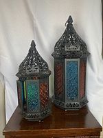 Photo showing front and angled view of both Moroccan style candle holders with colored glass panels and ornate metalwork, placed on a wooden cabinet.