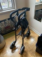 Photo of the folded Nexus walker showing handlebars with foam grips and the overall black metal frame, wheels, and folding mechanism.