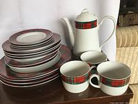 Photo of entire 17-piece white porcelain tea and dinner set with prominent red and green plaid patterns, showing teapot, cups, bowls, and plates stacked on a brown table.