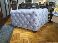 Tufted velvet ottoman cube in grey/silver displayed on hardwood floor, showing tufting and wooden legs.