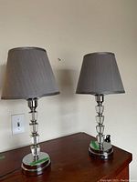 Two bedside lamps shown together on wooden table, with clear segmented crystal bases, chrome bottom plates, silver fabric shades. One shade shows slight opening.