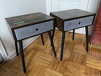 Two matching nightstands with dark wood particleboard tops and black legs, grey cloth basket drawer pulled closed.