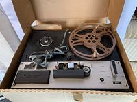 Full front-top view of the reel to reel tape recorder inside an open box showing controls and one tape reel with brown tape.