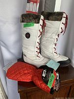Pair of white women's Cougar winter boots with red lace-up front, black soles, and faux fur trim, shown standing together next to a red ice scraper mitt.