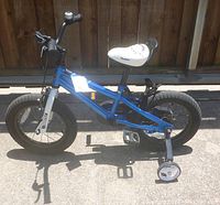Side view of the blue Royalbaby toddler bike with training wheels on pavement with wooden fence background.