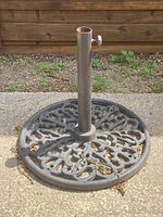 Cast iron umbrella stand photographed outdoors on concrete and gravel ground from a side angle showing base and central tube with tightening knob.