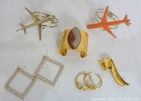 All six costume jewelry pieces displayed on white background including airplane bracelets, cuff bracelet with stone, rhinestone earrings, ram head earrings and ribbon brooch.