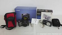 Canon PowerShot SX100 IS and Sony Cyber-shot DSC-W310 cameras with their original boxes and the red and black camera carrying case displayed on a table.