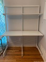 Front view of 48" wide freestanding Elfa desk system. Two white shelves mounted on vertical standards with metal legs and cross braces. Wooden floor and wall background.
