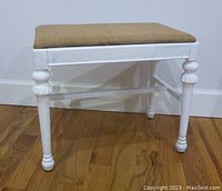 Front left angle view of the white farmhouse bench showing the burlap cushion, turned legs with decorative carved details, and cross supports.