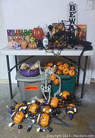 Photo showing a table with multiple Halloween pumpkin lights, spider decorations, skeleton cut-out, storage bins with Halloween labels, and various small pumpkin figurines arranged on and around the table.